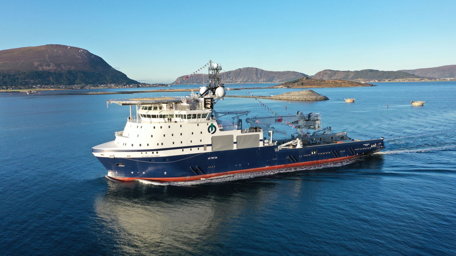 Prodotto - Offshore Tug/Supply Island Victory: Vista laterale della nave operativa in navigazione vicino alla costa, scafo blu e bianco, attrezzature avanzate e antenne sul ponte, sfondo con isole e colline.
