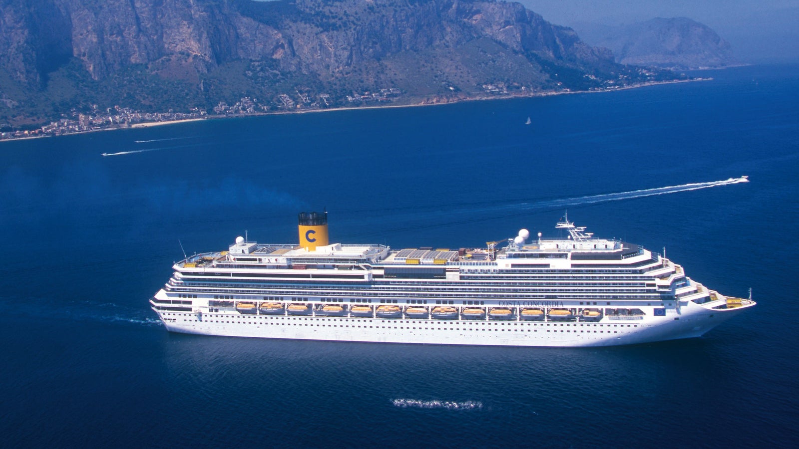 Fincantieri | Group - Business - Products - Cruise Ships - Costa Concordia: View of Costa Concordia cruise ship in operation navigating near the coast, white hull with multiple passenger decks, yellow funnel with C logo, mountainous background.