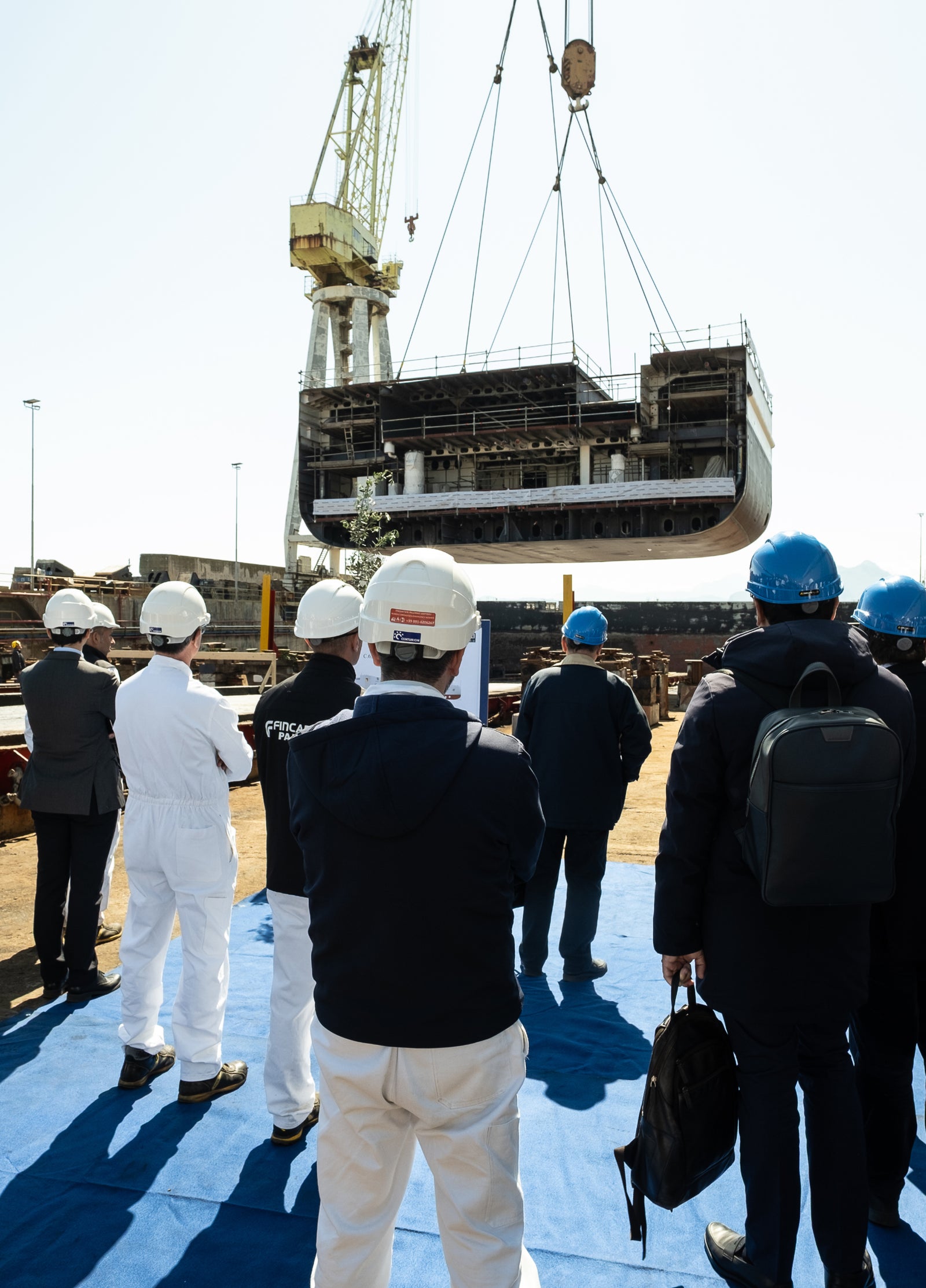  Fincantieri | Eccellenza industriale - Sostenibilità - Operazioni di cantiere: Gruppo di lavoratori con caschi e divise osserva il sollevamento di una grande sezione di scafo nave tramite gru, attrezzature industriali e cielo sereno sullo sfondo. Ambiente professionale orientato alla sicurezza.