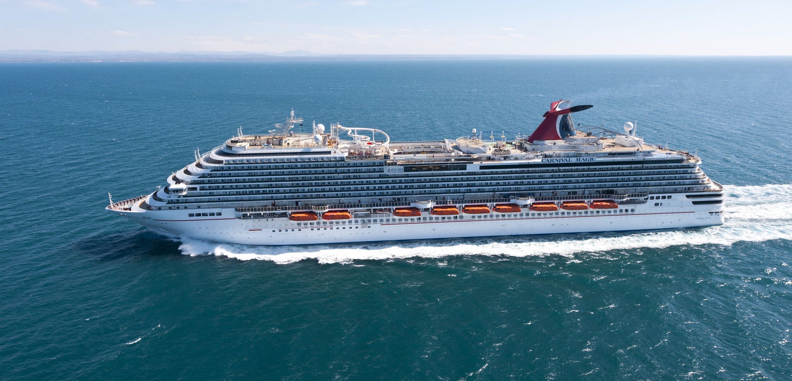 Fincantieri | Group - Business - Products - Cruise Ships - Carnival Magic: Large operational white cruise ship navigating calm open sea under clear blue sky, multiple decks, lifeboats and distinctive red funnel, tranquil waters and wide horizon emphasize scale and peaceful setting.