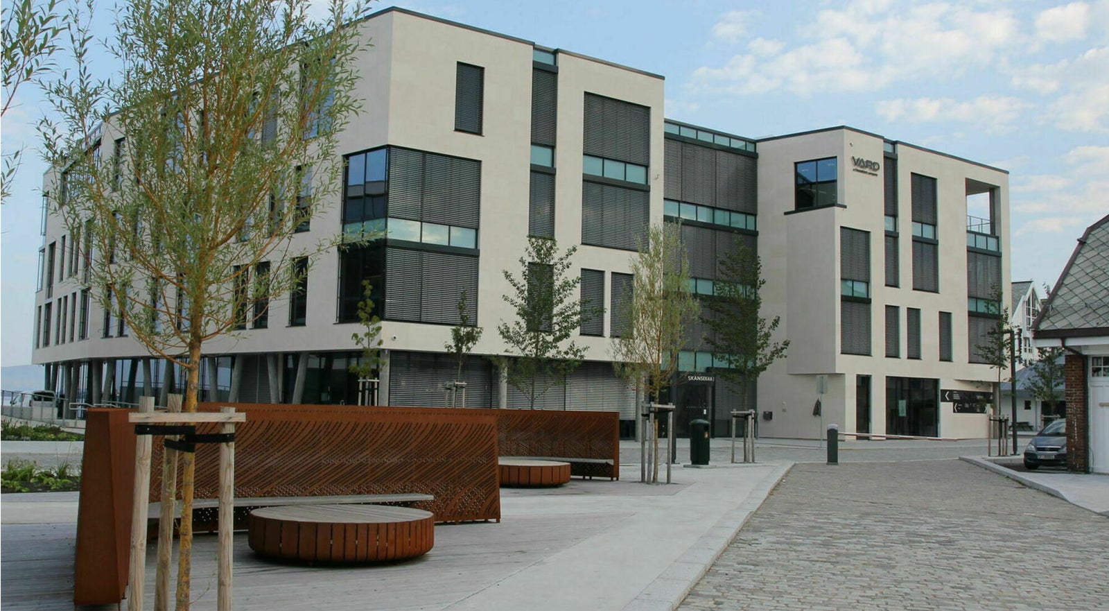 A contemporary multi-story office building with large glass windows and a beige facade. The setting features a landscaped outdoor area with benches, trees, and paved walkways under a partly cloudy sky. The building includes visible signage reading 'HUBRO' on the upper corner.
