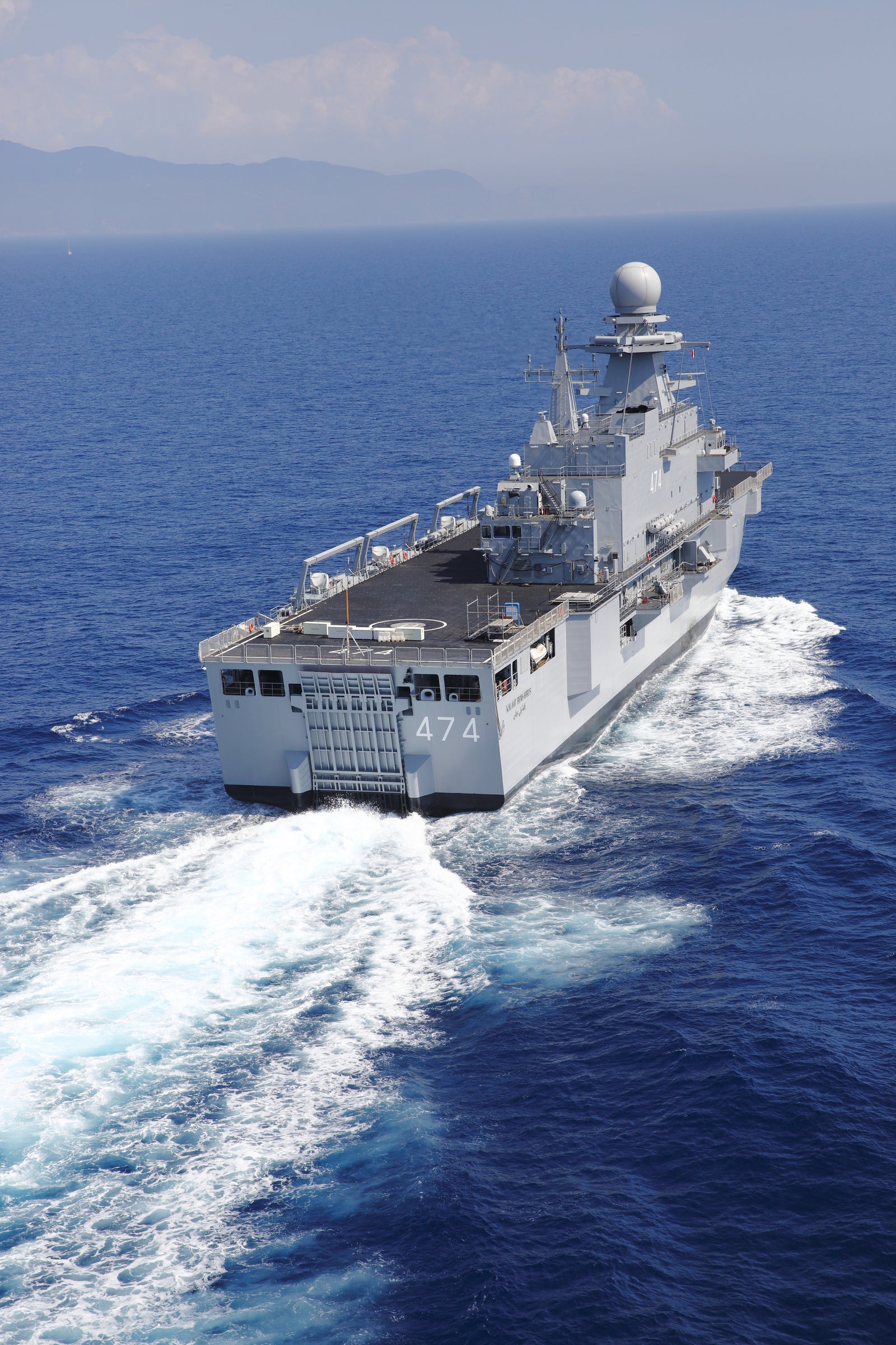 Fincantieri | Gruppo - Business - Prodotti - Navi Militari - Portaerei e Navi Anfibi - Kalaat Beni Abbes: Grande nave militare "Kalaat Beni Abbes" in operazione, in navigazione su acque blu, scia bianca visibile, montagne in lontananza sull'orizzonte.