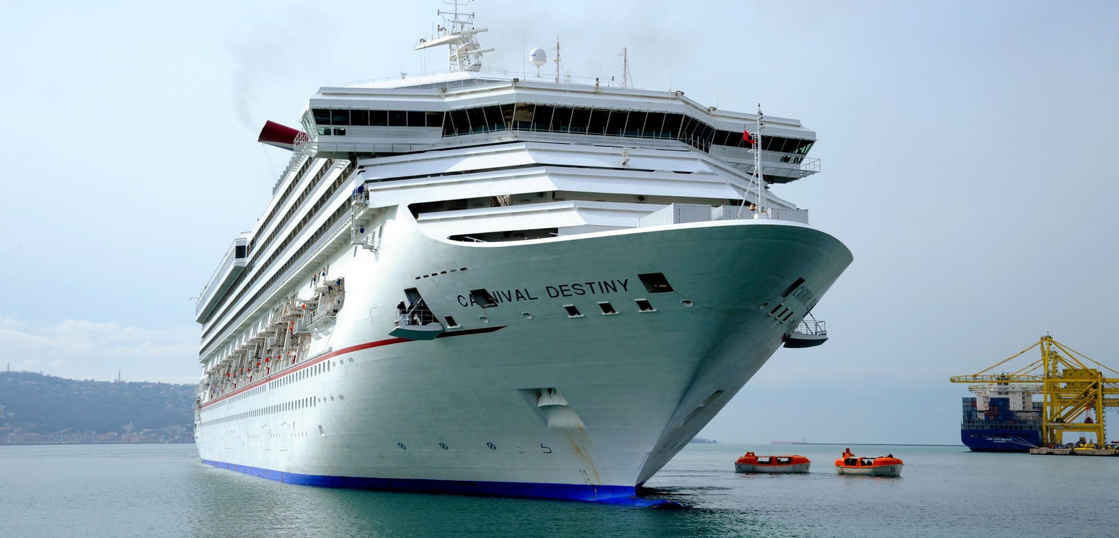 Fincantieri | Group - Business - Products - Cruise Ships - Carnival Destiny: Bow view of large white cruise ship 'Carnival Destiny' docked in port, white exterior with red and blue accents, surrounded by calm waters, yellow crane and industrial structures in background, ship name clearly shown on side highlighting its identity.