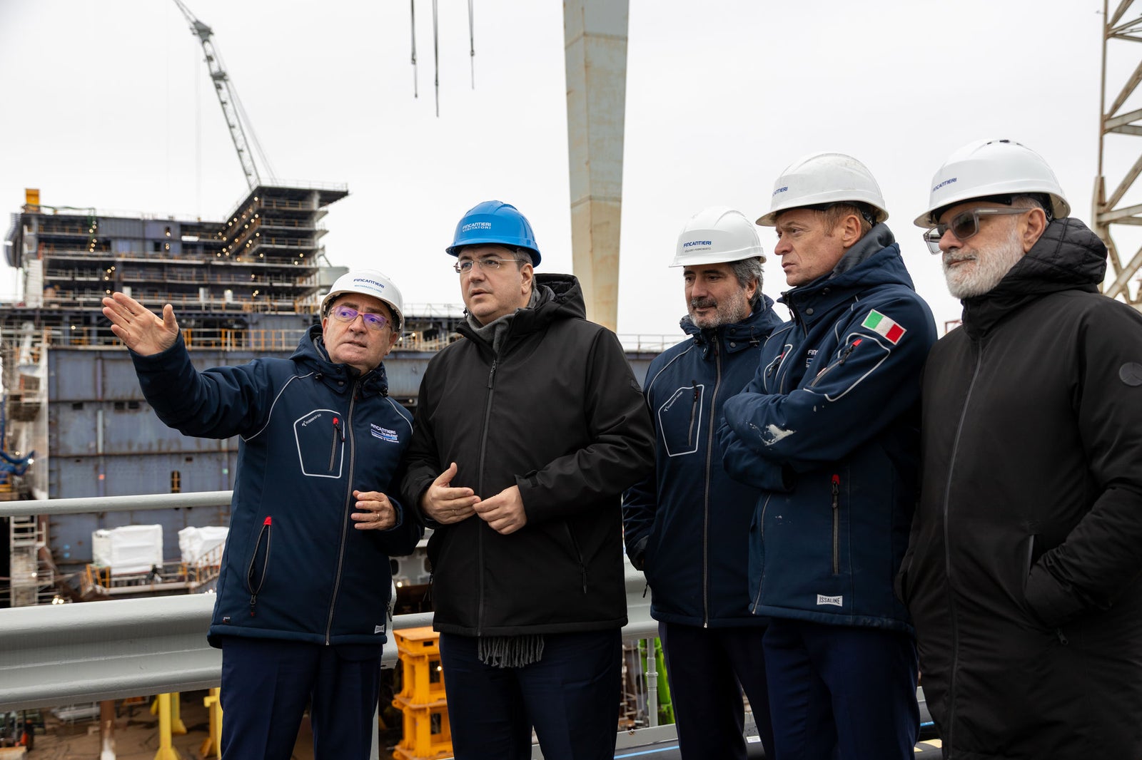 Newsroom - Comunicati stampa - 2025 - Gruppo Fincantieri: Fincantieri: visita del Commissario Europeo Apostolos Tzitzikostas al cantiere di Monfalcone: Il Commissario Europeo Apostolos Tzitzikostas con tecnici e manager Fincantieri al cantiere navale di Monfalcone, indossano caschi di sicurezza e abbigliamento tecnico, struttura navale in costruzione e gru sullo sfondo.