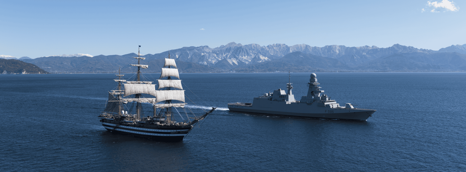 Fincantieri | About Us - Heritage and Future: Historic sailing ship with sails unfurled and modern warship operating on calm blue waters, mountainous background, contrast between naval tradition and innovation under clear sky and daylight.