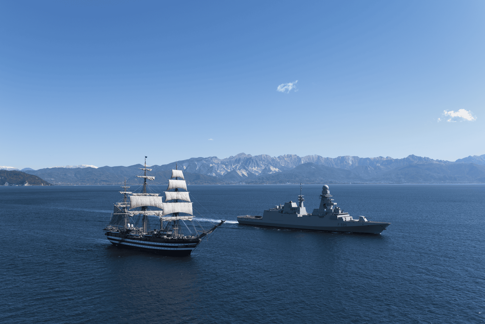 Fincantieri | Chi siamo - Eredità e Futuro: Veliero storico a vele spiegate e nave da guerra moderna operano su acque blu calme, sfondo montuoso, contrasto tra tradizione e innovazione navale sotto cielo sereno e illuminazione diurna.