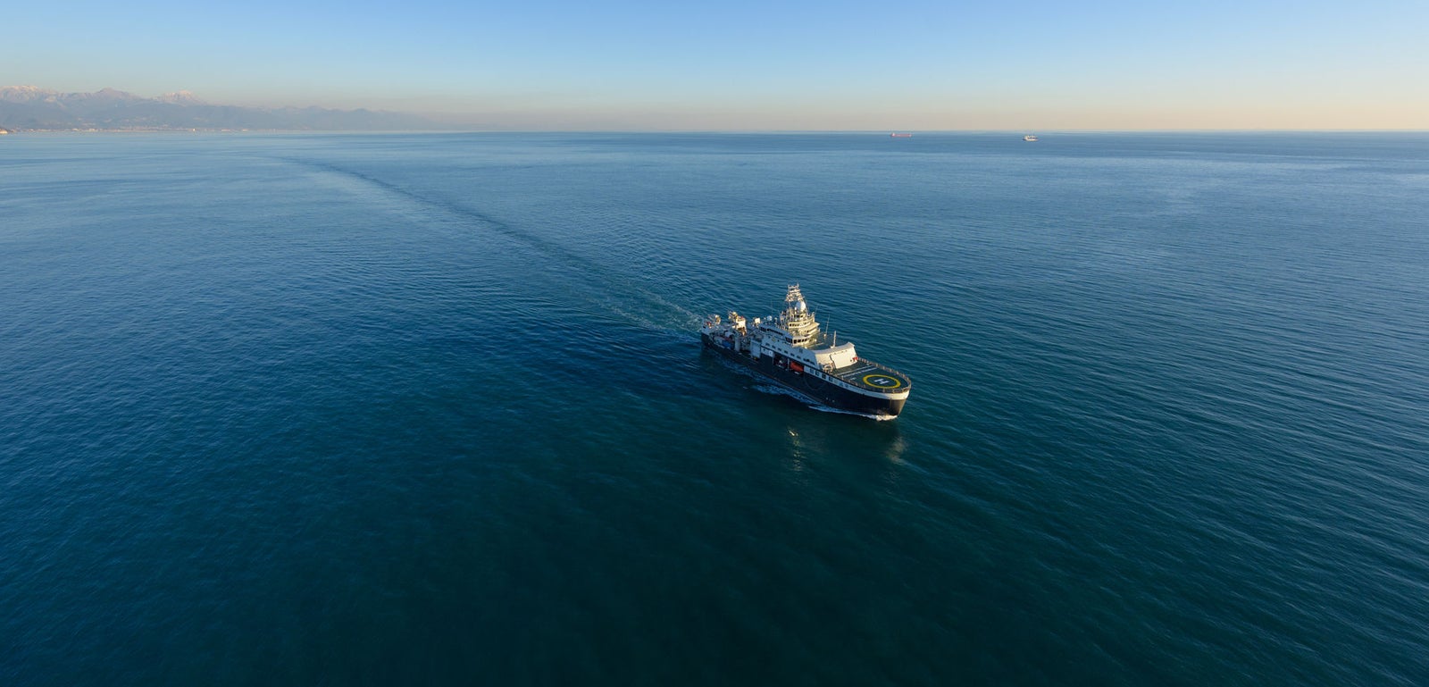 Prodotto - Nave da Ricerca Polare: Vista laterale della nave operativa in navigazione, scia visibile sull’acqua, sfondo con orizzonte ampio e profili montuosi distanti.