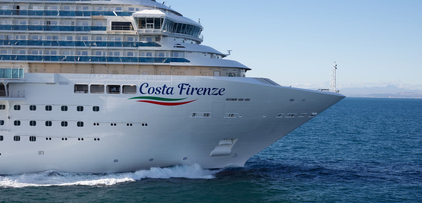  Fincantieri | Group - Business - Products - Cruise Ships - Costa Firenze: Costa Firenze cruise ship in operation sailing through calm blue waters, sleek white exterior and multiple decks, name 'Costa Firenze' and wave logo visible on side, clear horizon with distant lighthouse in background.