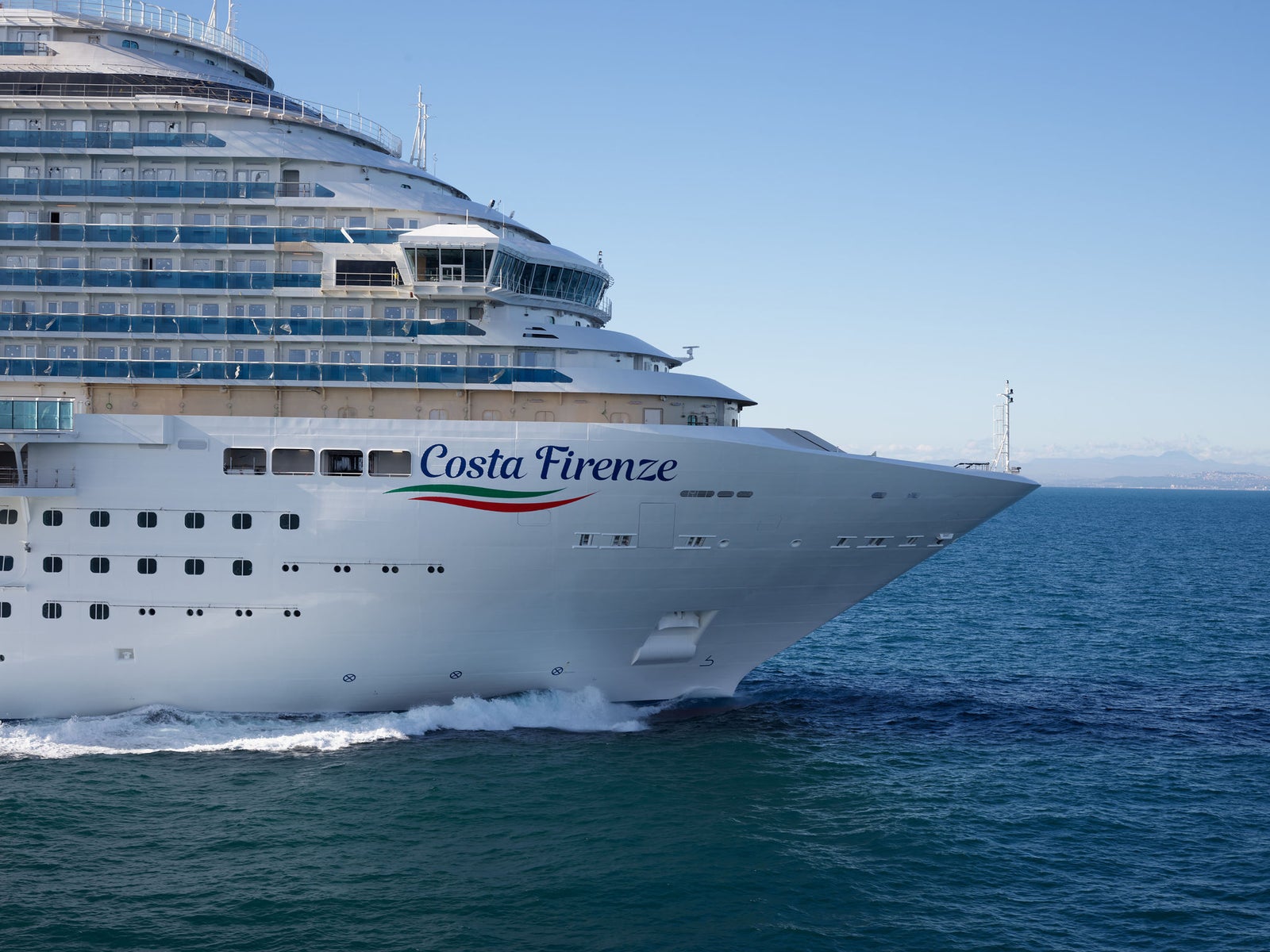  Fincantieri | Group - Business - Products - Cruise Ships - Costa Firenze: Costa Firenze cruise ship in operation sailing through calm blue waters, sleek white exterior and multiple decks, name 'Costa Firenze' and wave logo visible on side, clear horizon with distant lighthouse in background.