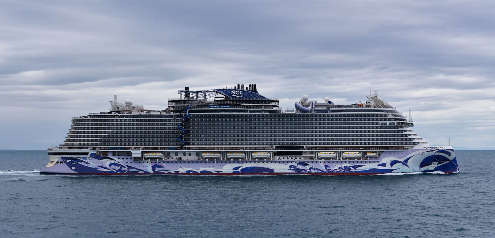 Fincantieri | Gruppo - Business - Prodotti - Navi da Crociera - Norwegian Viva: Nave da crociera moderna operativa con design intricati blu e bianco, naviga su acque aperte calme sotto cielo nuvoloso, più ponti e comfort visibili, scenario sereno ed espansivo.