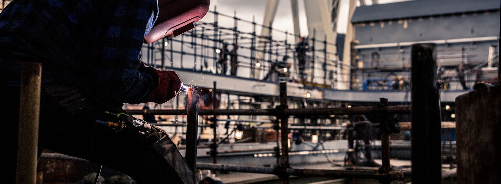 Fincantieri | Business Services - After-Sales Services - Lifecycle Management: A welder wearing protective gear is focused on welding metal at a shipyard. The setting features scaffolding, industrial structures, and a large ship in the background. The scene is under a covered area with dramatic lighting and cloudy skies, emphasizing the industrial mood.