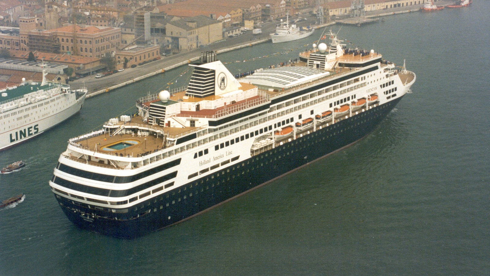 Fincantieri | Group - Business - Products - Cruise Ships - Veendam: Side view of Veendam cruise ship docked at port, multiple decks, visible pool, distinctive white-black exterior design, historic architecture in background, presence of smaller boats and second ship.