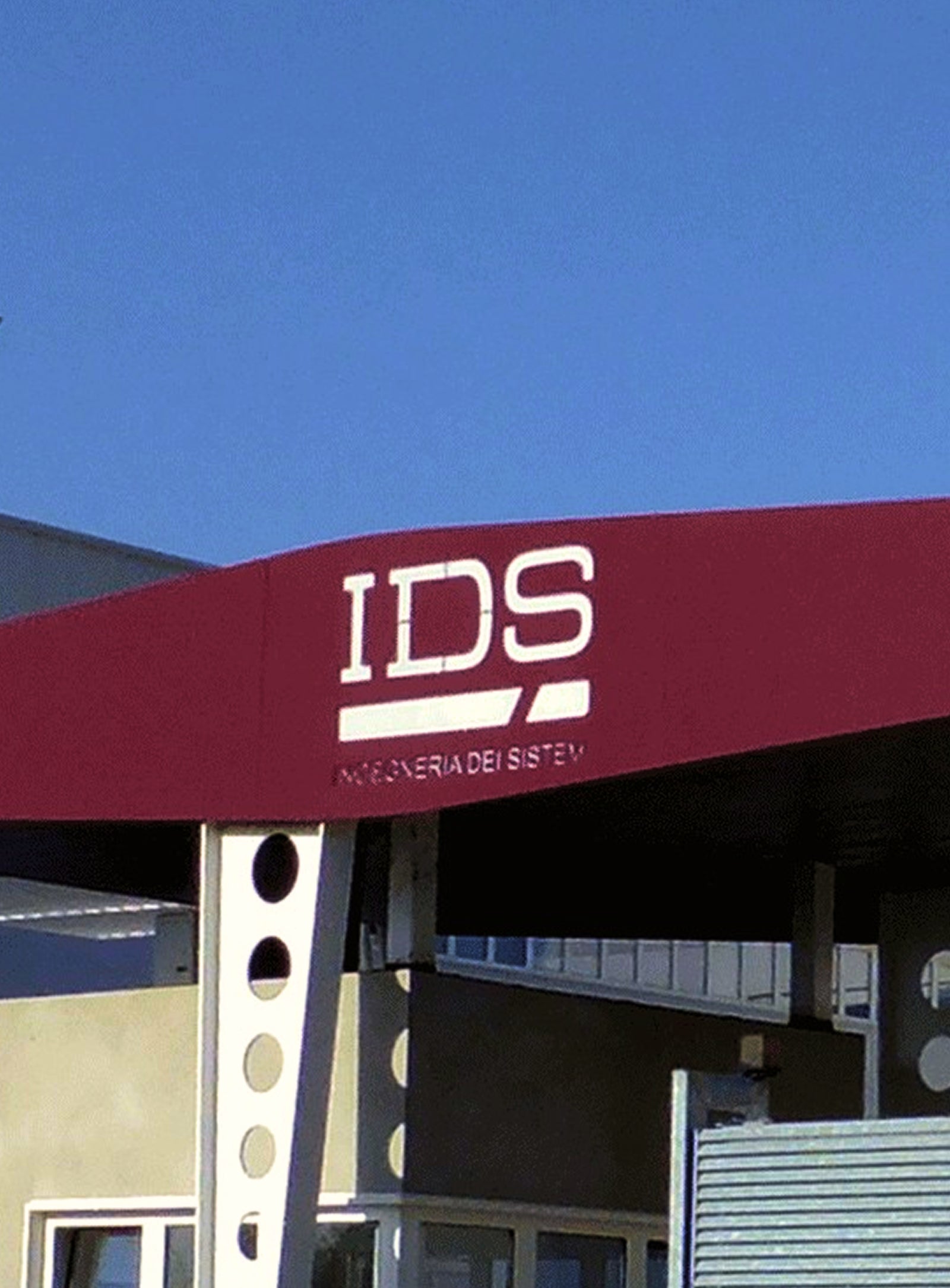  Fincantieri | Gruppo - Società - Controllate e collegate - IDS Ingegneria di sistemi: Pensilina d’ingresso di edificio dal design architettonico moderno, testo ‘IDS’ in bianco su sfondo marrone; contesto con edifici industriali e cielo blu sereno per ambiente professionale e pulito.