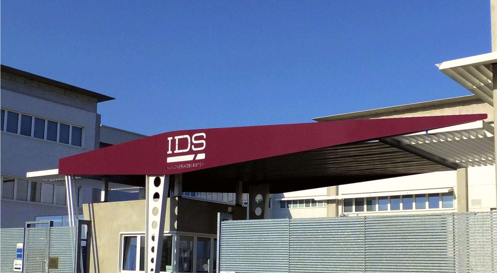 Fincantieri | Gruppo - Società - Controllate e collegate - IDS Ingegneria di sistemi: Pensilina d’ingresso di edificio dal design architettonico moderno, testo ‘IDS’ in bianco su sfondo marrone; contesto con edifici industriali e cielo blu sereno per ambiente professionale e pulito.