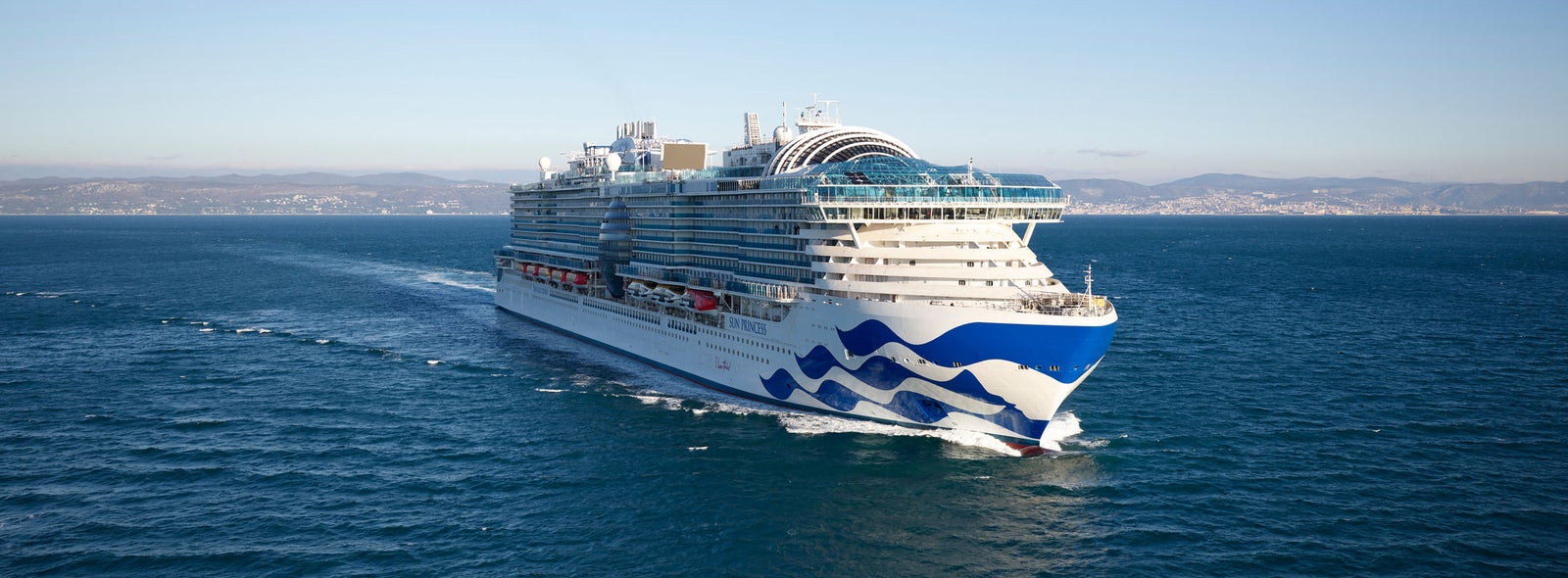 Fincantieri | Group - Innovation - Naval Ecosystem of the Future - Propulsion Systems and Clean Fuels: Large operational cruise ship sailing on calm waters, clear blue sky, hull with wave pattern and multiple decks with visible amenities, coastal land in the background.