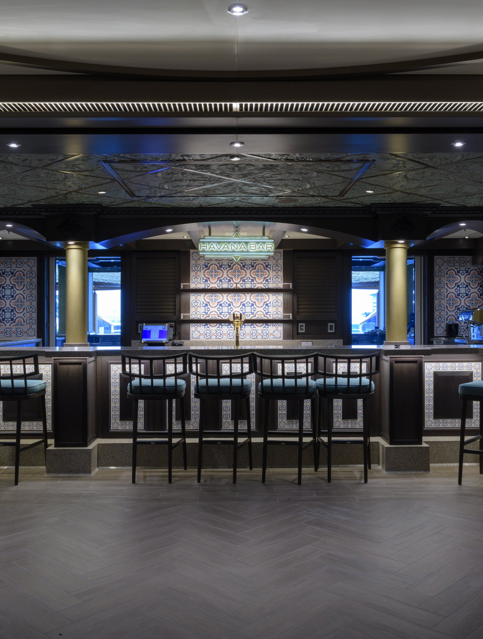 Fincantieri | Group - Business - Products - Cruise Ships - Carnival Horizon: Elegant bar counter with tall stools in center, surrounded by intricate tiled motifs and warm lighting, modern and luxurious environment, spacious layout and ceiling decorative details, scene without people highlighting architectural and design elements.