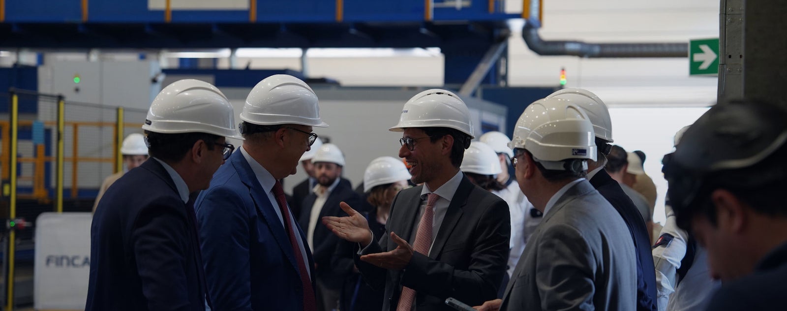Fincantieri | Gruppo - Sostenibilità - Eccellenza industriale - Cilenti: Gruppo di persone con caschi di sicurezza bianchi e abiti formali riunite in ambiente di fabbrica industriale, macchinari, segnaletica di sicurezza e attrezzature visibili nello sfondo; contesto di ispezione o discussione professionale in stabilimento produttivo.