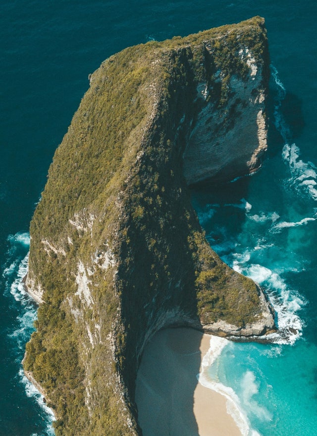 Fincantieri | Group - Sustainability - Industrial Excellence - Products: Aerial photo of a majestic rock formation surrounded by turquoise ocean waters, isolated beach at the base with lush vegetation and rocks, vivid colors and natural details.