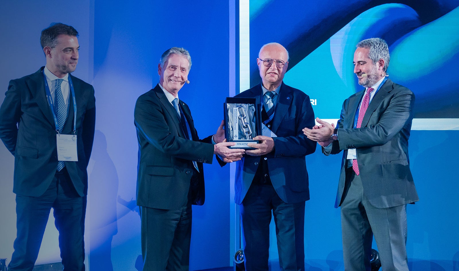 Fornitori e Partner – Fincantieri | Partnership: Gruppo di uomini in abiti formali su un palco durante una cerimonia di premiazione, uno tiene una targa o trofeo mentre gli altri applaudono, sfondo blu con visual astratte, ambiente professionale e corporate. 2° Fincantieri's Supplier Summit