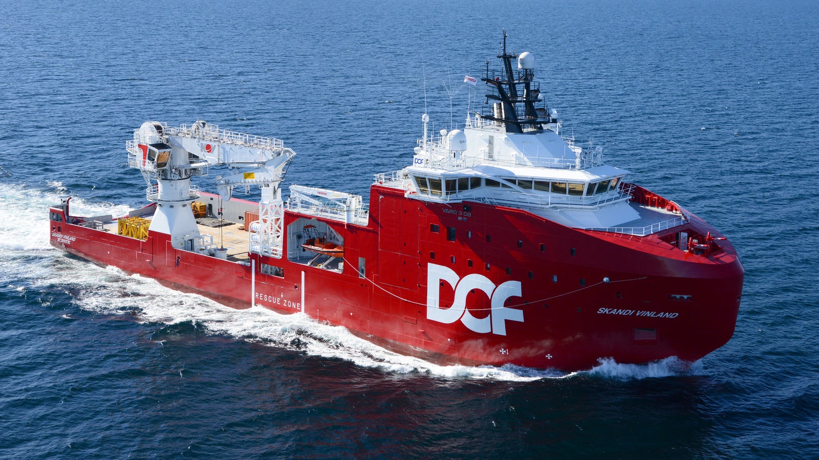 Fincantieri | Group - Business - Products - Offshore and Specialized Vessels - Energy - OFFSHORE SUBSEA CONSTRUCTIOn VESSEL - Skandi Vinland: Striking red "Skandi Vinland" offshore vessel with "DOF" branding in operation, advanced crane and deck machinery, navigating through fjord, mountainous terrain, clear blue water, bright sky.