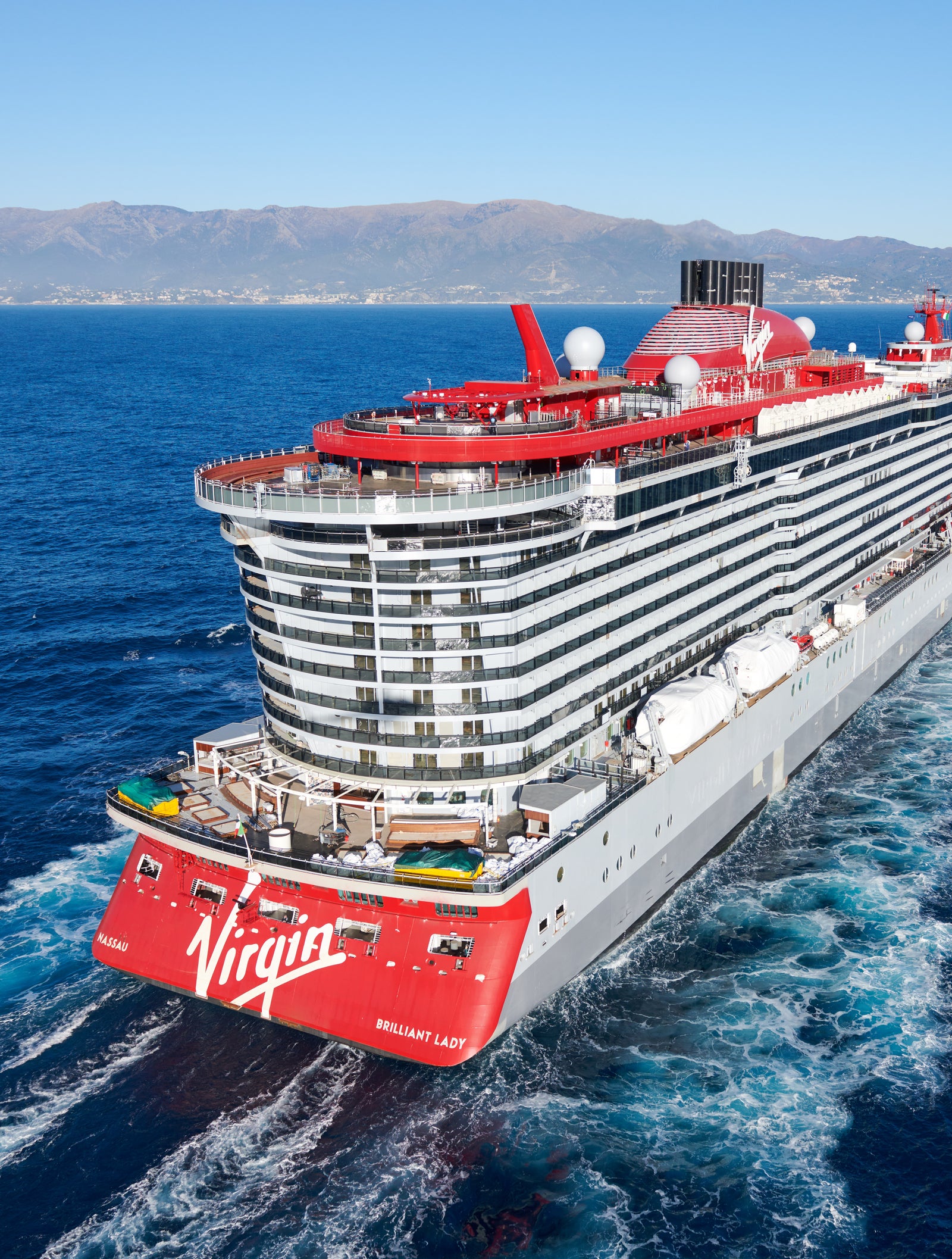 Fincantieri | Gruppo - Business - Prodotti - Navi da Crociera - Brilliant Lady: Grande nave Virgin Voyages operativa con design elegante e dettagli rossi, molti ponti e cabine di vari tipi, nome 'Brilliant Lady' visibile sullo scafo, naviga su acque calme sotto cielo parzialmente nuvoloso.