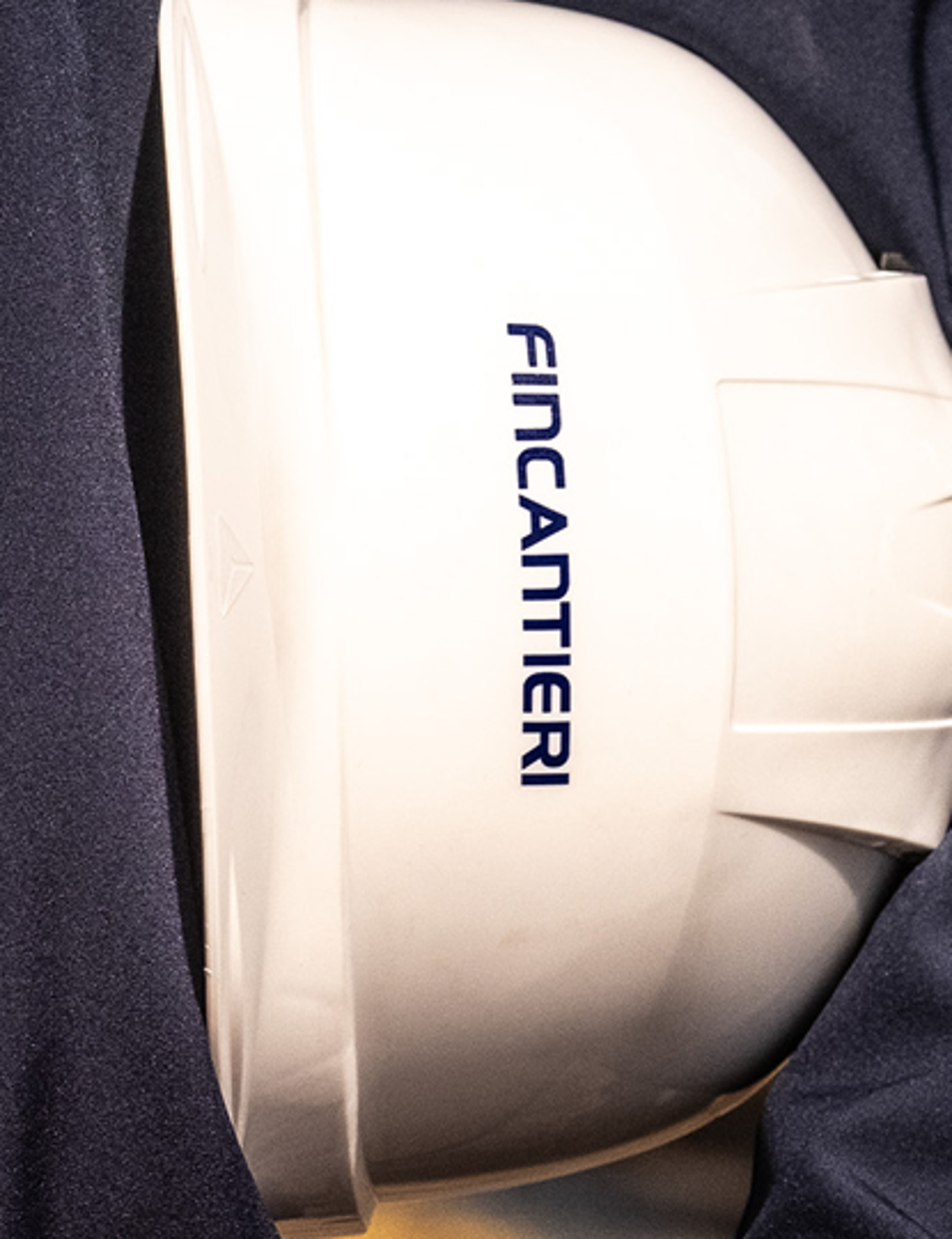Fincantieri | Persone - Inclusione Vivere Fincantieri: Primo piano di persona in giacca blu che regge casco bianco con logo Fincantieri ben visibile, contesto esterno in area di cantiere o industriale, focus su mani e casco. Programmi formativiione Vivere Fincantieri: Primo piano di persona in giacca blu che regge casco bianco con logo Fincantieri ben visibile, contesto esterno in area di cantiere o industriale, focus su mani e casco.