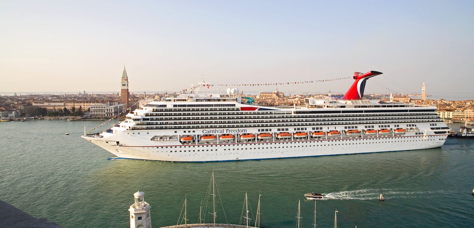  Fincantieri | Gruppo - Business - Prodotti - Navi da Crociera - Carnival Freedom: Grande nave da crociera operativa che scivola attraverso il porto di Venezia, esterno bianco con accenti rossi e neri, iconico fumaiolo ben visibile, barche più piccole e architettura storica veneziana, incluso un campanile prominente, acque calme e cielo limpido valorizzano l’ambientazione.