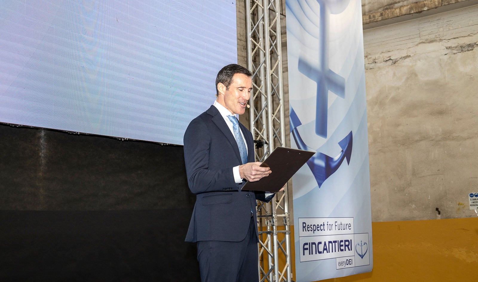 Fornitori e Partner – Fincantieri | Partnership: Uomo in abito formale sul palco con clipboard durante una presentazione, sfondo con grande schermo e banner con logo Fincantieri, testo “Respect for Future”, ancora nautica e scritta “Fincantieri S.p.A.”, presenza di elementi industriali e illuminazione scenica, 2° Supplier Summit Fincantieri.