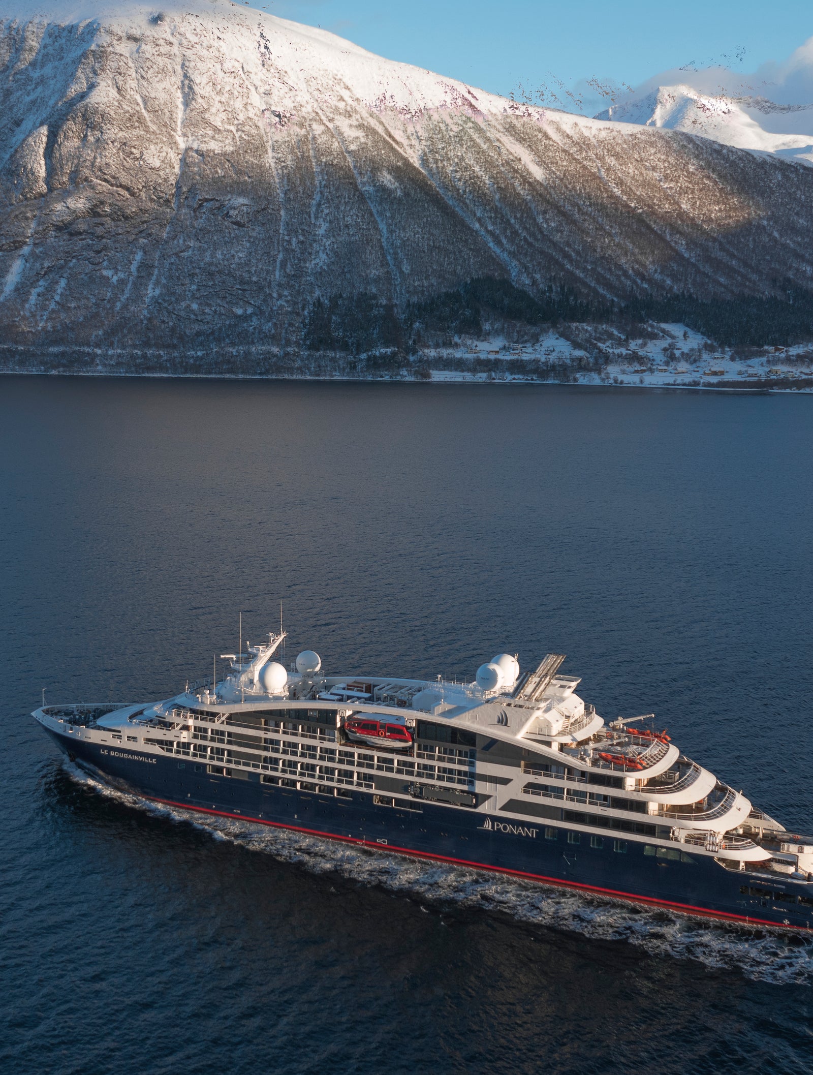  Fincantieri | Group - Business - Products - Cruise Ships - Le Bougainville: Modern luxury cruise ship in operation sailing through serene fjord surrounded by snow-capped mountains, sleek design with visible red and blue accents, calm waters and dramatic landscapes under clear sky.