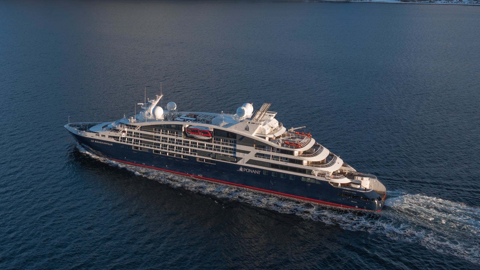 Fincantieri | Gruppo - Business - Prodotti - Navi da Crociera - Le Bougainville: Vista laterale della nave Le Bougainville operativa in navigazione all’interno di un fiordo, circondata da montagne innevate e acque calme che riflettono il paesaggio, scafo dal profilo lineare con dettagli rossi e blu visibili.