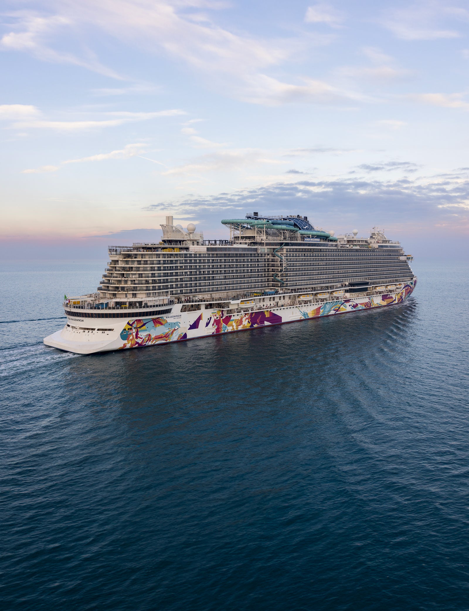 Fincantieri | Group - About Us - Strategic Vision - Business Model: A large, modern cruise ship adorned with vibrant artistic designs sails on calm ocean waters during sunset. The vessel features multiple decks and expansive amenities, with clear skies and soft clouds in the background. The serene setting highlights the grandeur and leisure of ocean travel.