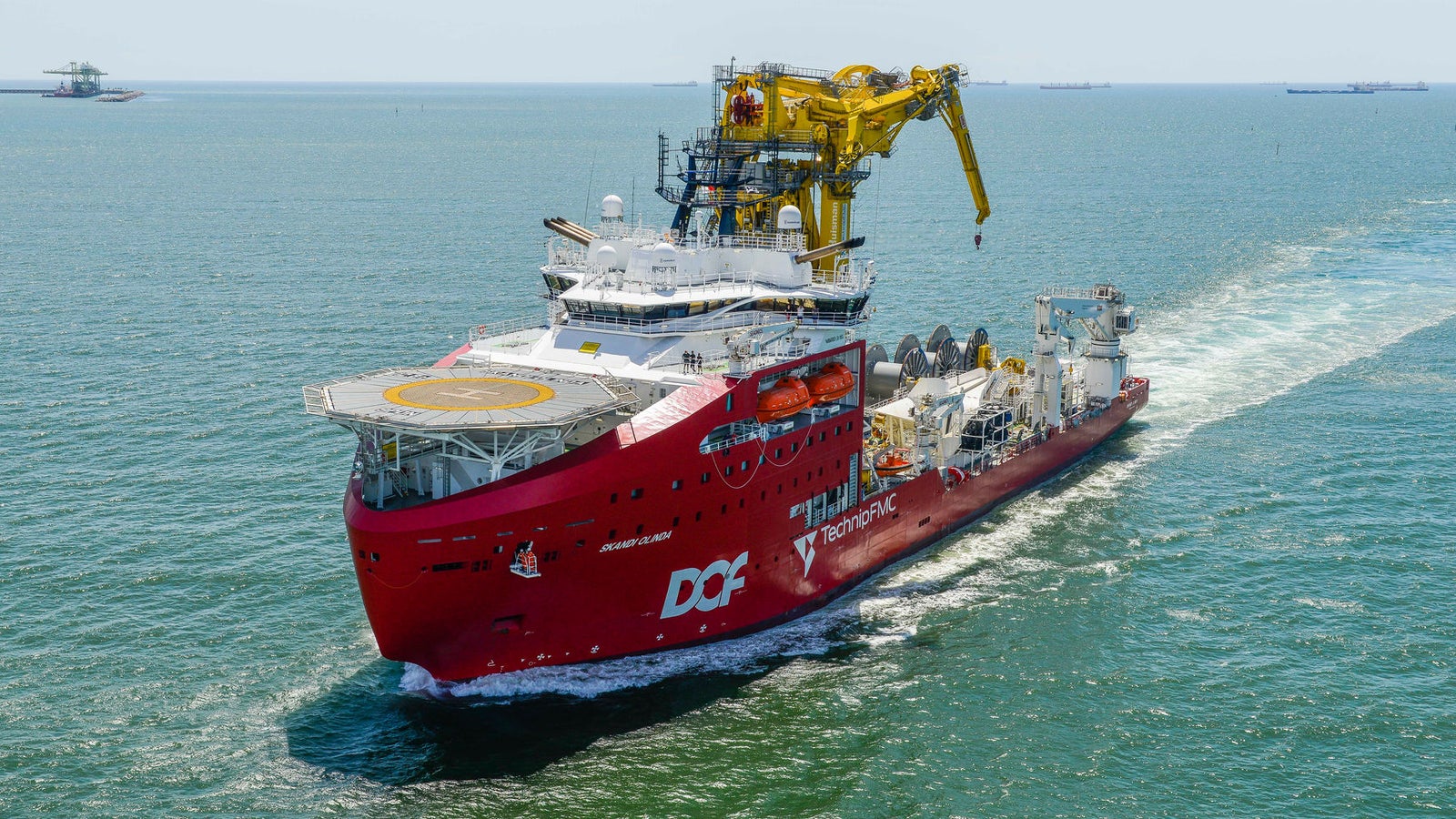 Prodotto - Offshore Support Vessel Skandi Onlinda: Vista laterale della nave operativa in navigazione, scafo rosso, gru gialla, scritte DOF e TechnipFMC ben visibili, sfondo con strutture industriali distanti.