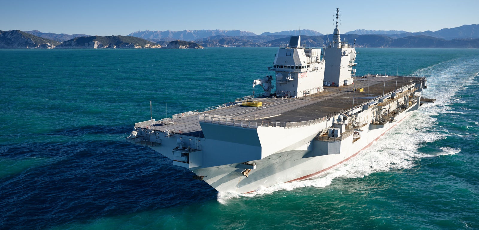Fincantieri | Group - Business - Products - Naval Vessels - Aircraft Carriers and Amphibious Ships - Trieste: Large gray aircraft carrier "Trieste" in operation, sailing through turquoise water, clear blue sky, visible white wake.