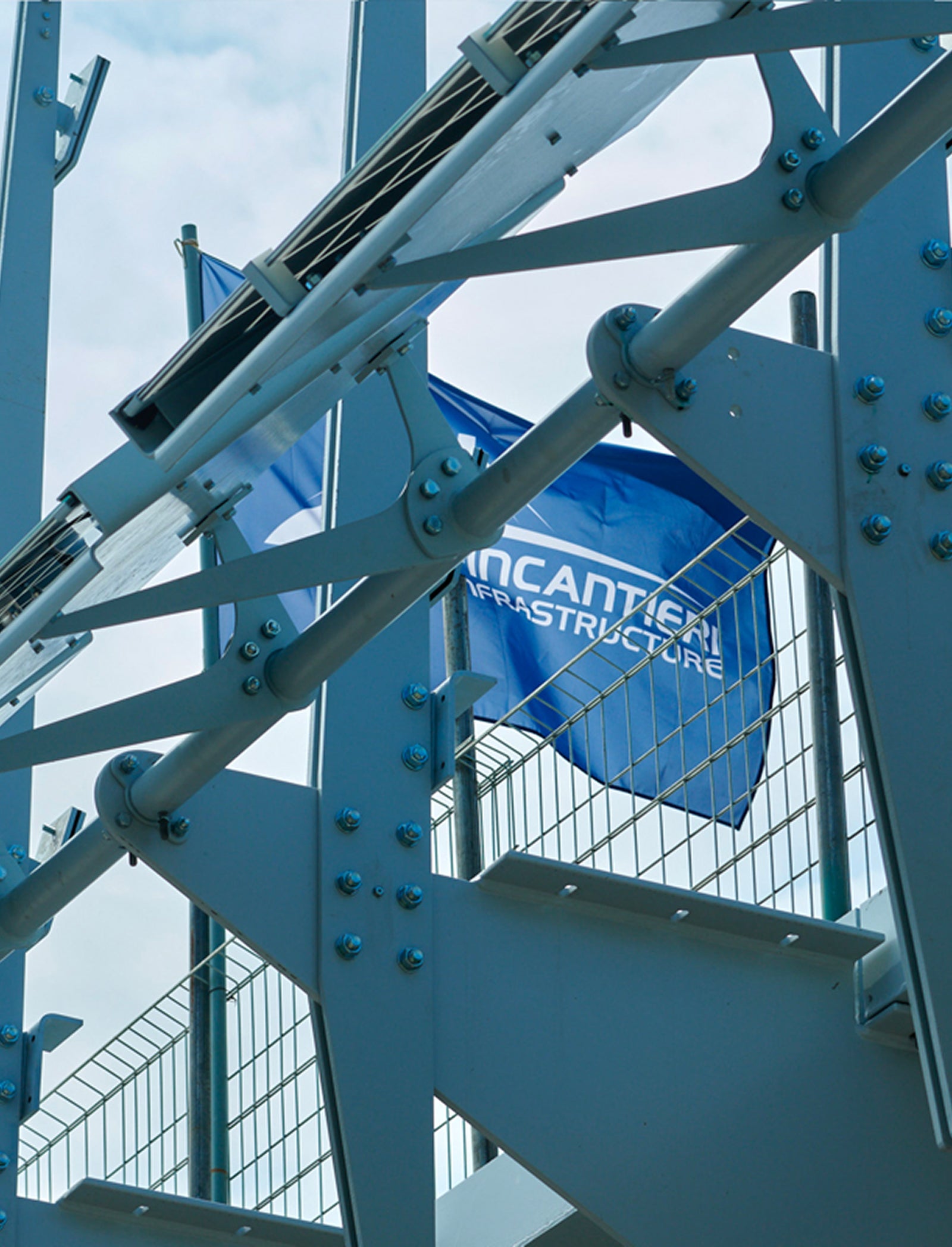 Fincantieri | Group - Subsidiaries and Affiliates - Fincantieri Infrastructure: Close-up view of blue industrial structure featuring metal beams and bolts; flag with ‘Fincantieri Infrastructure’ text visible in the background, geometric design and sturdy construction under partly cloudy sky.