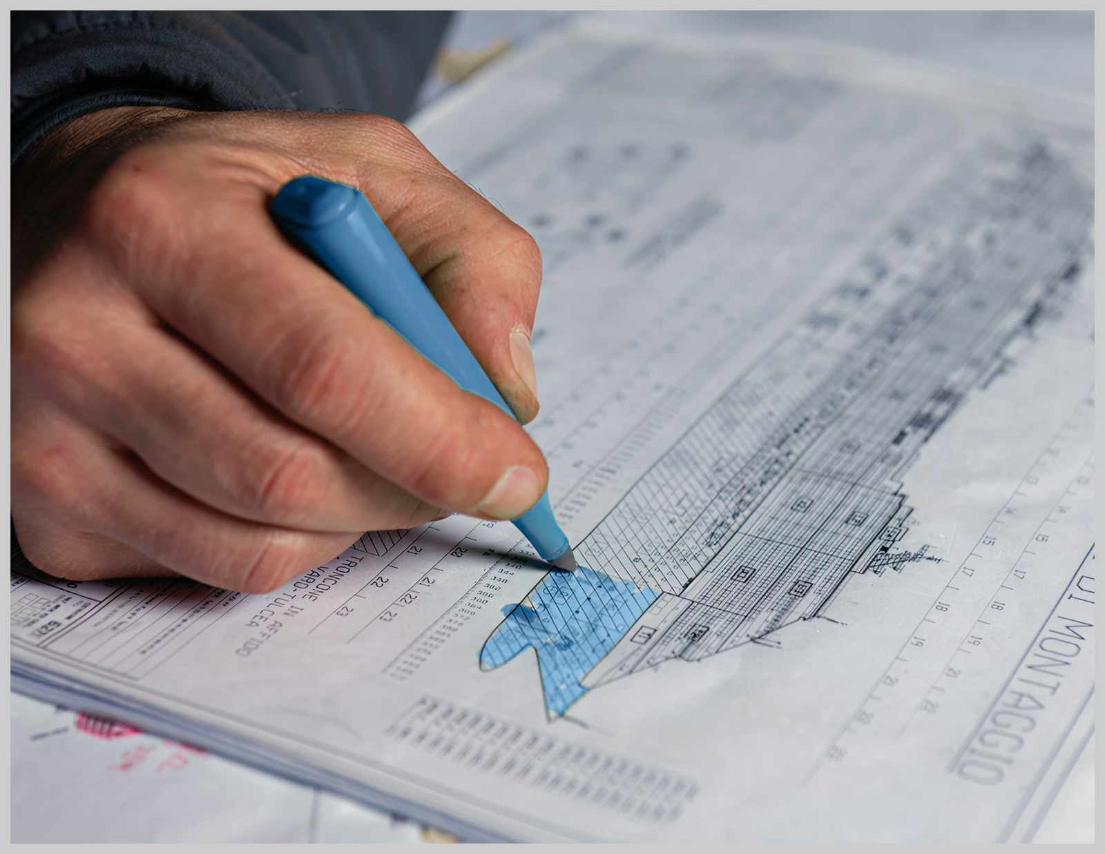 Fincantieri | People - Diversity, Equity and Inclusion: Close-up of a hand coloring a section of a naval blueprint with a blue marker, technical details clearly visible, workspace or design studio setting, focus on precision and structural detail.