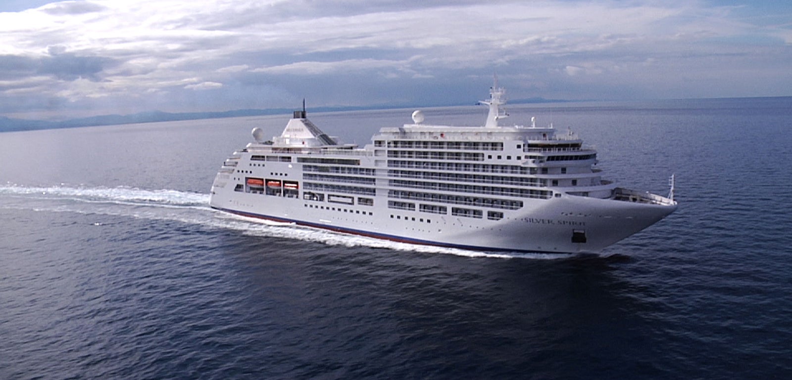 Fincantieri | Group - Business - Products - Cruise Ships - Silver Spirit: Large modern operational white cruise ship with multiple decks and visible lifeboats, sailing on calm waters under partly cloudy sky, visible wake, atmosphere of luxury, safety, and relaxation, highlighting the ship’s elegance and grandeur.