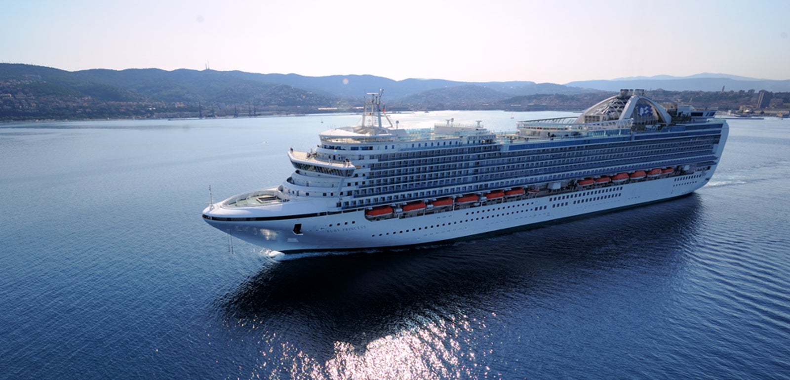 Fincantieri | Gruppo - Business - Prodotti - Navi da Crociera - Ruby Princess: Nave da crociera imponente operativa che scivola su acque calme e aperte sotto cielo azzurro limpido, più ponti, scialuppe di salvataggio, colline panoramiche visibili in lontananza.