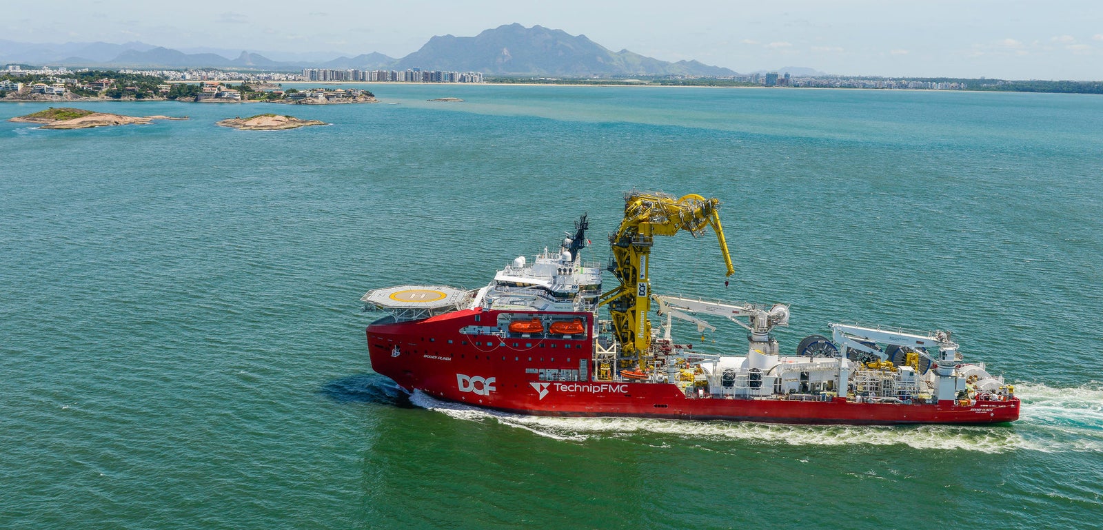 Prodotto - Offshore Support Vessel Skandi Onlinda: Vista laterale della nave operativa in navigazione, scafo rosso, grande gru gialla, scritte DOF e TechnipFMC visibili, sfondo città costiera e catena montuosa.