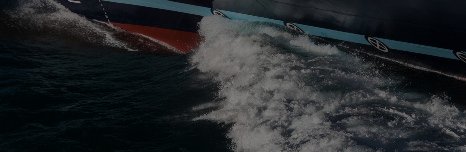 Fincantieri | Group - Business - Products - Systems Integration - Ship in operation: Large ship cuts through dark, choppy water, creating a significant wake; hull painted red and dark blue, visible as it moves forward.