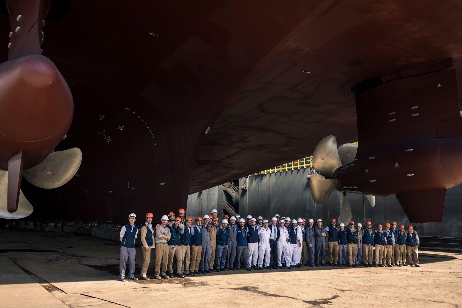 Fincantieri – Investing in Fincantieri: Low-angle view of a large ship in a dry dock, with propellers and rudders clearly visible; a group of people in uniforms and helmets stands under the hull, industrial setting, sunlight illuminating parts of the hull.