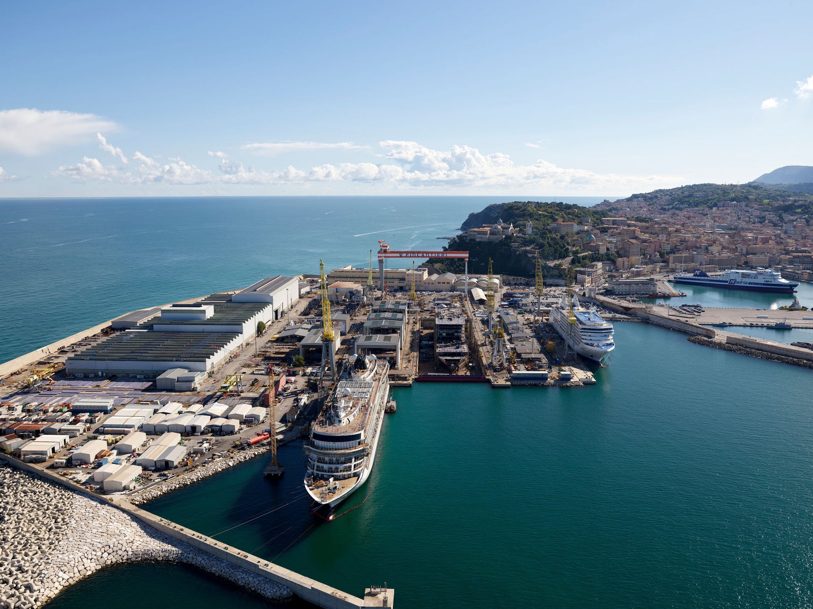 Fincantieri | Products - Systems, Components and Infrastructure - Shipyard: Large coastal facility with ships under construction and moored, industrial cranes and warehouses; coastal city visible in the background, clear sky.