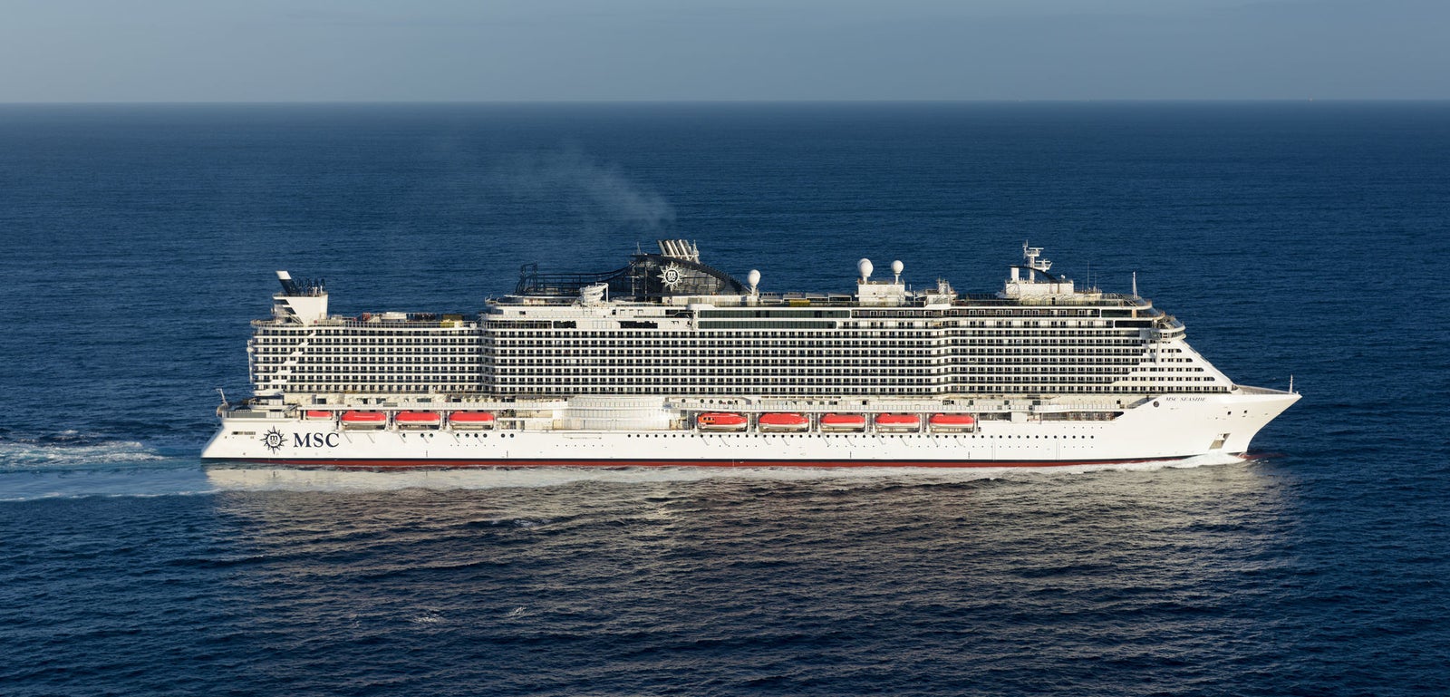 Fincantieri | Group - Business - Products - Cruise Ships - MSC Seaside: Side view of MSC Seaside cruise ship in operation, massive hull with visible passenger decks, red lifeboats, smoke from funnel, MSC logo on hull.