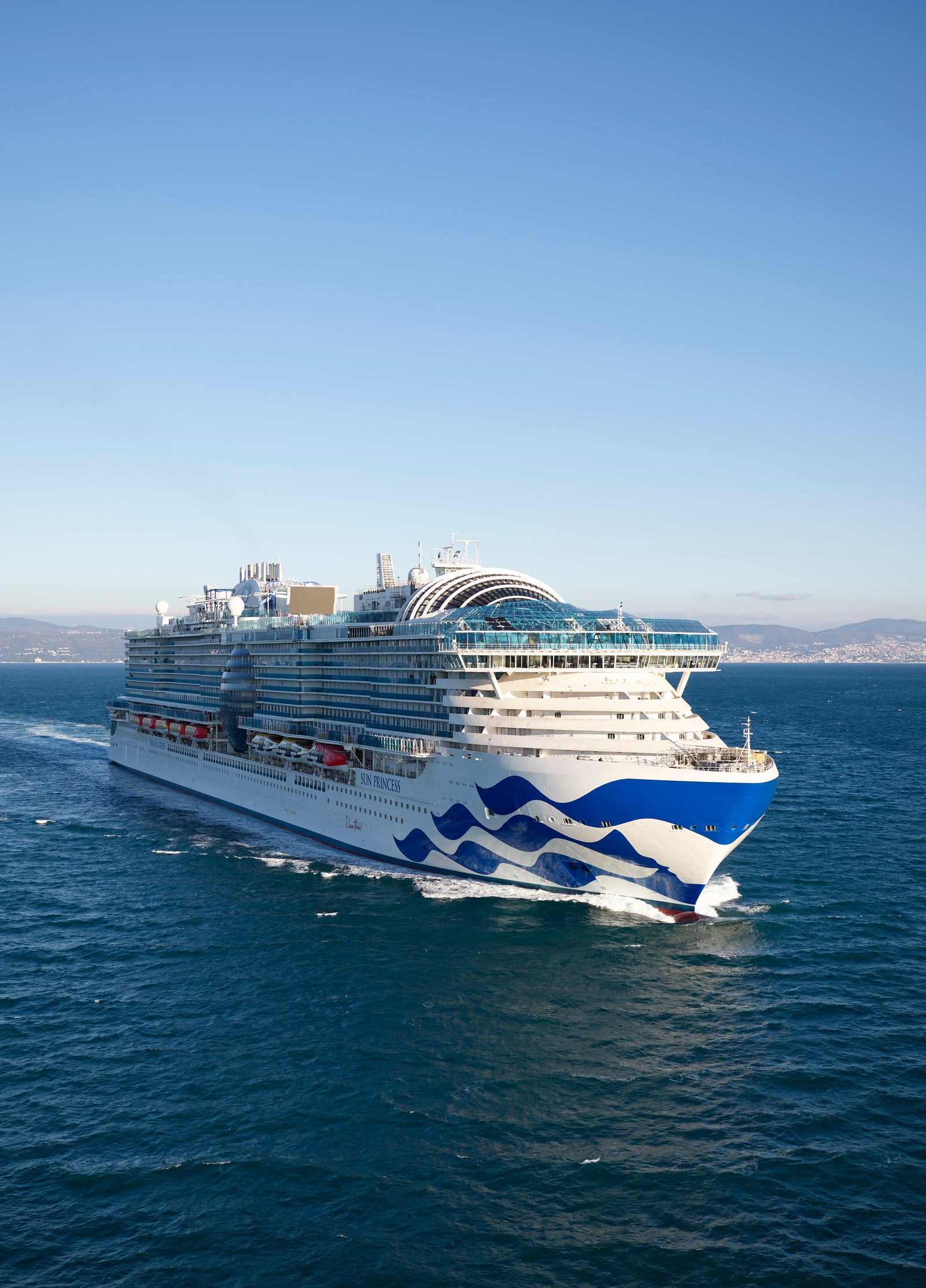 Fincantieri Group | About Us: Side view of a large cruise ship in operation, sailing on calm waters, featuring a distinctive wave pattern on the hull, multiple visible decks and amenities, with a distant coastline and clear sky in the background.