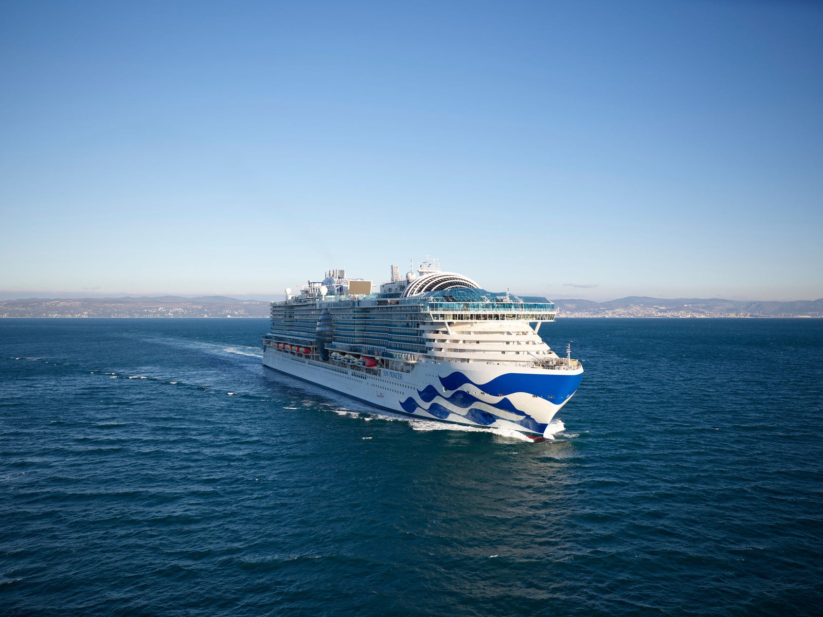 Gruppo Fincantieri | Chi siamo: Vista laterale di una grande nave da crociera operativa in navigazione su acque calme, scafo con motivo a onda distintivo, multipli ponti e servizi visibili, sfondo con costa lontana e cielo sereno.