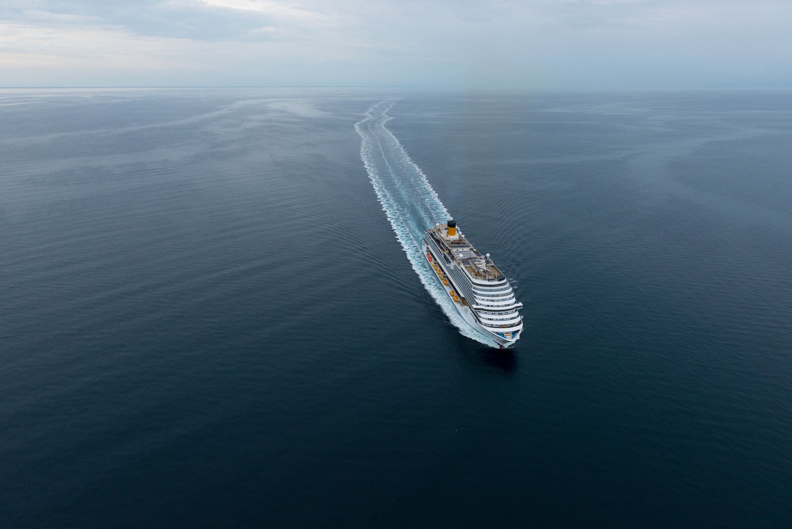 Fincantieri | Group - Business - Products - Cruise Ships - Costa Diadema: Large cruise ship in operation navigating calm open ocean waters, visible wake behind, overcast skies and expansive sea views, multiple decks and vibrant accents emphasize grandeur and scale.