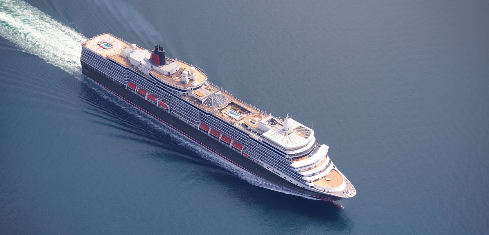Fincantieri | Group - Business - Products - Cruise Ships - Queen Elizabeth: Aerial view of Queen Elizabeth cruise ship in operation navigating open sea, well-defined wake in water, multiple passenger decks with pool area and visible outdoor spaces, linear profile hull and distinctive funnel.