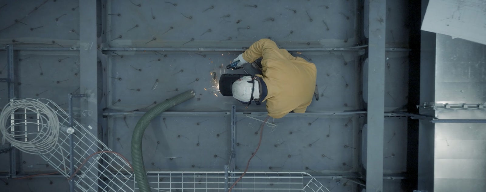Fincantieri | Group - Sustainability - Industrial Excellence - Health and Safety: Group of people wearing orange safety helmets and blue shirts, gathered in a construction site or outdoor industrial location with scaffolding in the background, one person with a smartwatch, the shirts display the text "Pinnacle... Explore". Team atmosphere and professional readiness.