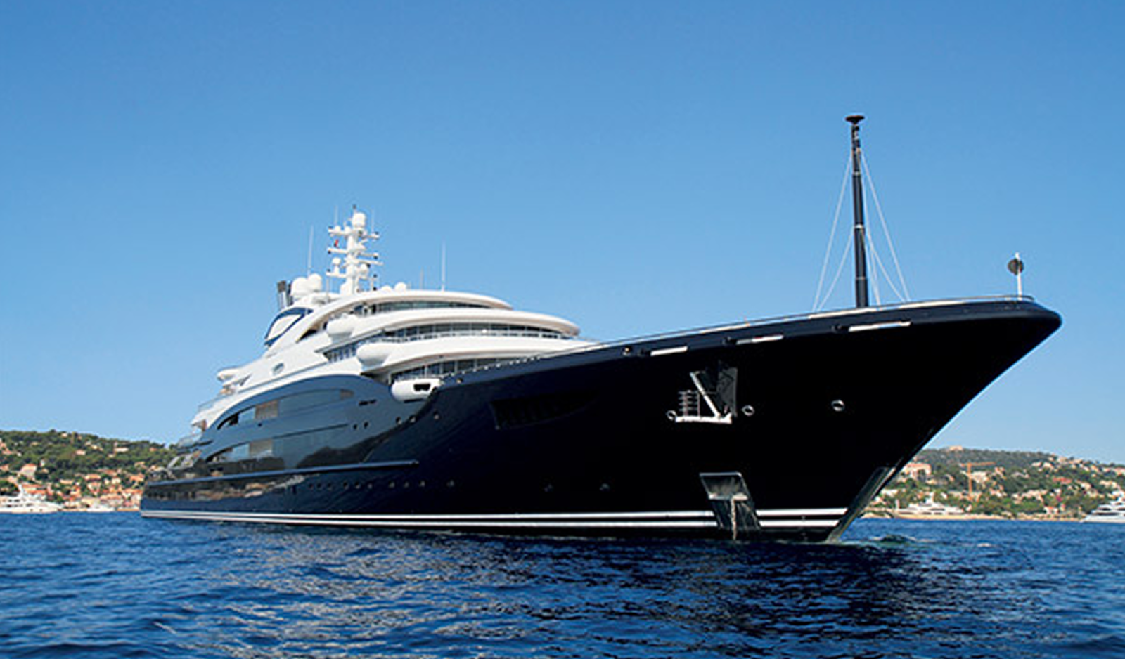 Fincantieri | About Us - Heritage and Future: Modern luxury yacht with a sleek appearance, glossy black hull, and multiple decks, floating on calm blue waters, scenic coastal background in the distance, clear sky.