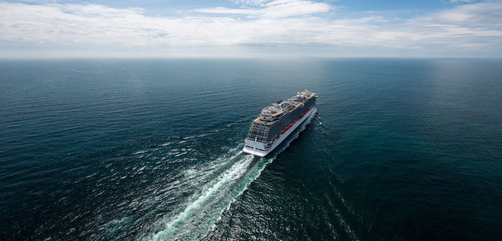 Fincantieri | Group - Business - Products - Cruise Ships - Royal Princess: Stern view of large luxury cruise ship in operation sailing on calm open waters under partly cloudy sky, visible wake emphasizing movement.