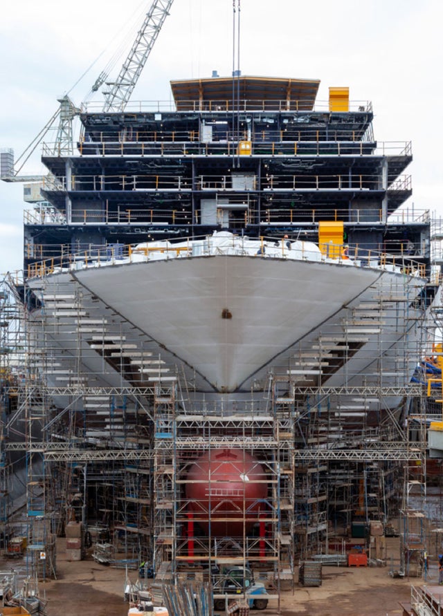 Fincantieri | Group - Sustainability - Energy Transition - Net-zero: Large ship under construction in a dry dock, surrounded by scaffolding and industrial equipment, shipyard with cranes and workers in the background; bow in the foreground highlights scale and engineering complexity.
